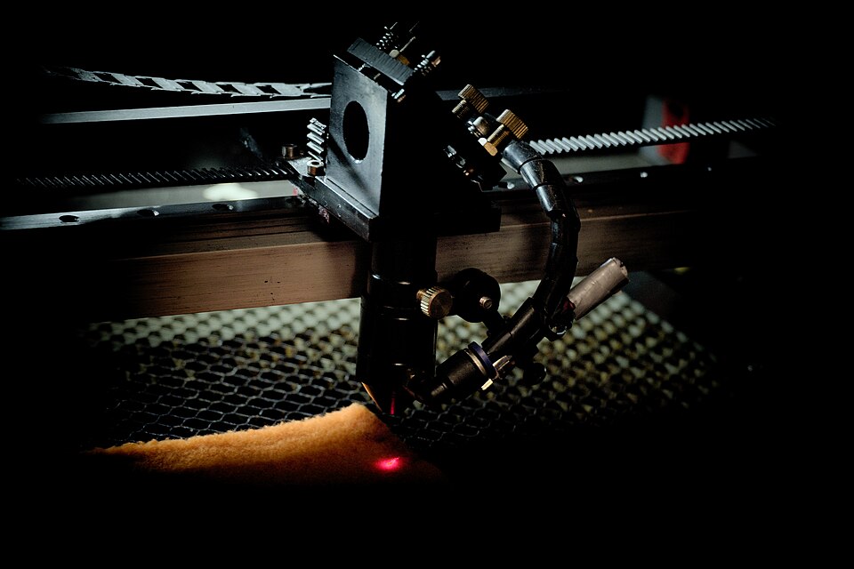 Figure 4 — A laser cutting head of the general type: it carries the final mirror, the focusing lens, and the air-assist nozzle, and moves along the gantry above the work. Source: Wikimedia Commons …