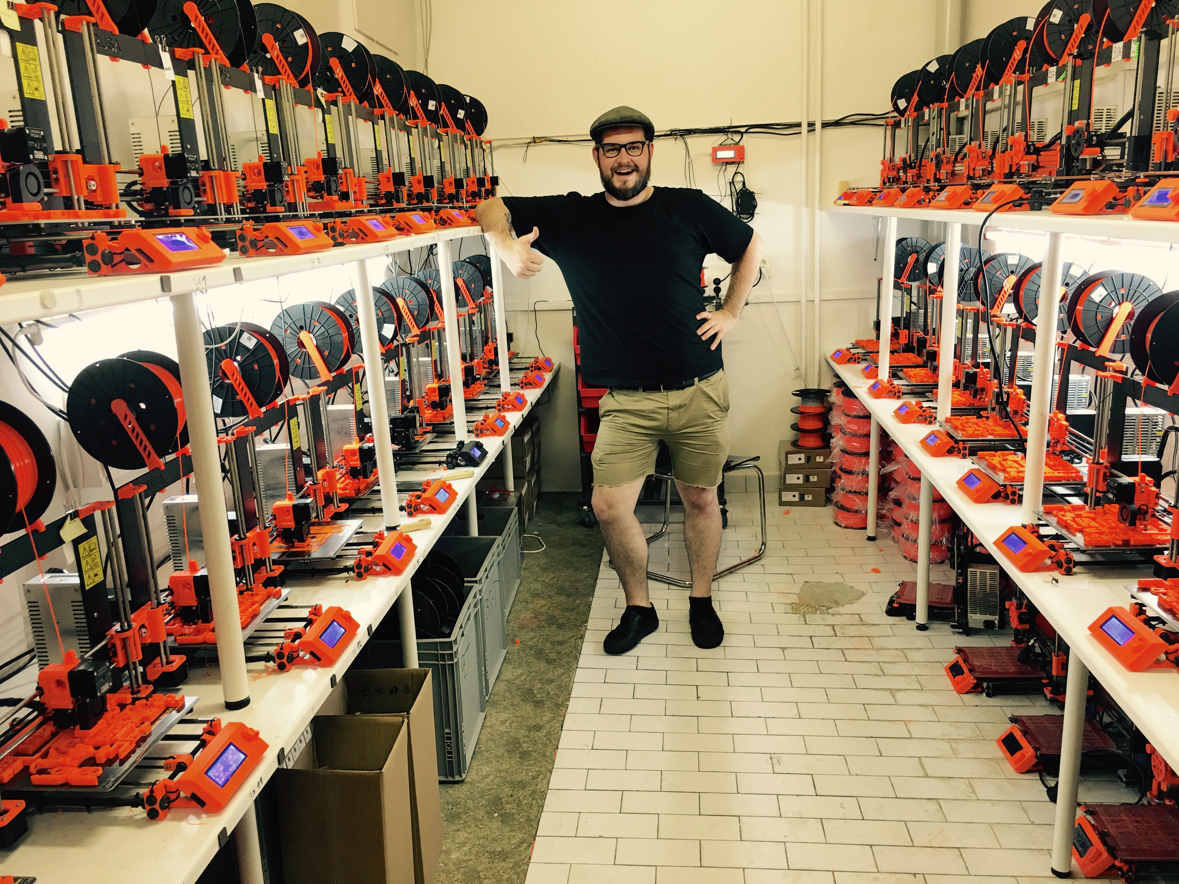 Figure 3 — Prusa Research's own print farm: rows of identical Original Prusa machines producing the printed parts for new printers. A design that behaves identically across dozens of units, run aro…